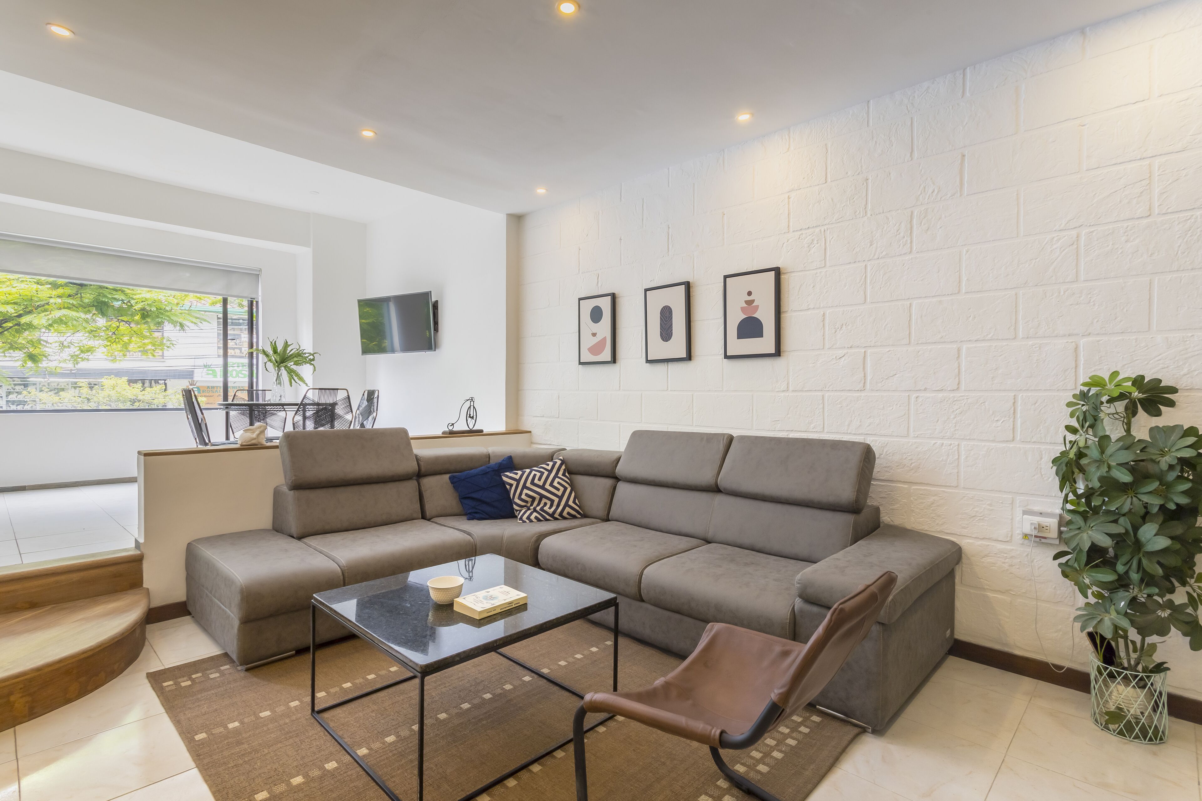 A modern living room with a sectional sofa, a glass coffee table, and a brown leather chair. There is a large window providing natural light and a view of greenery outside.