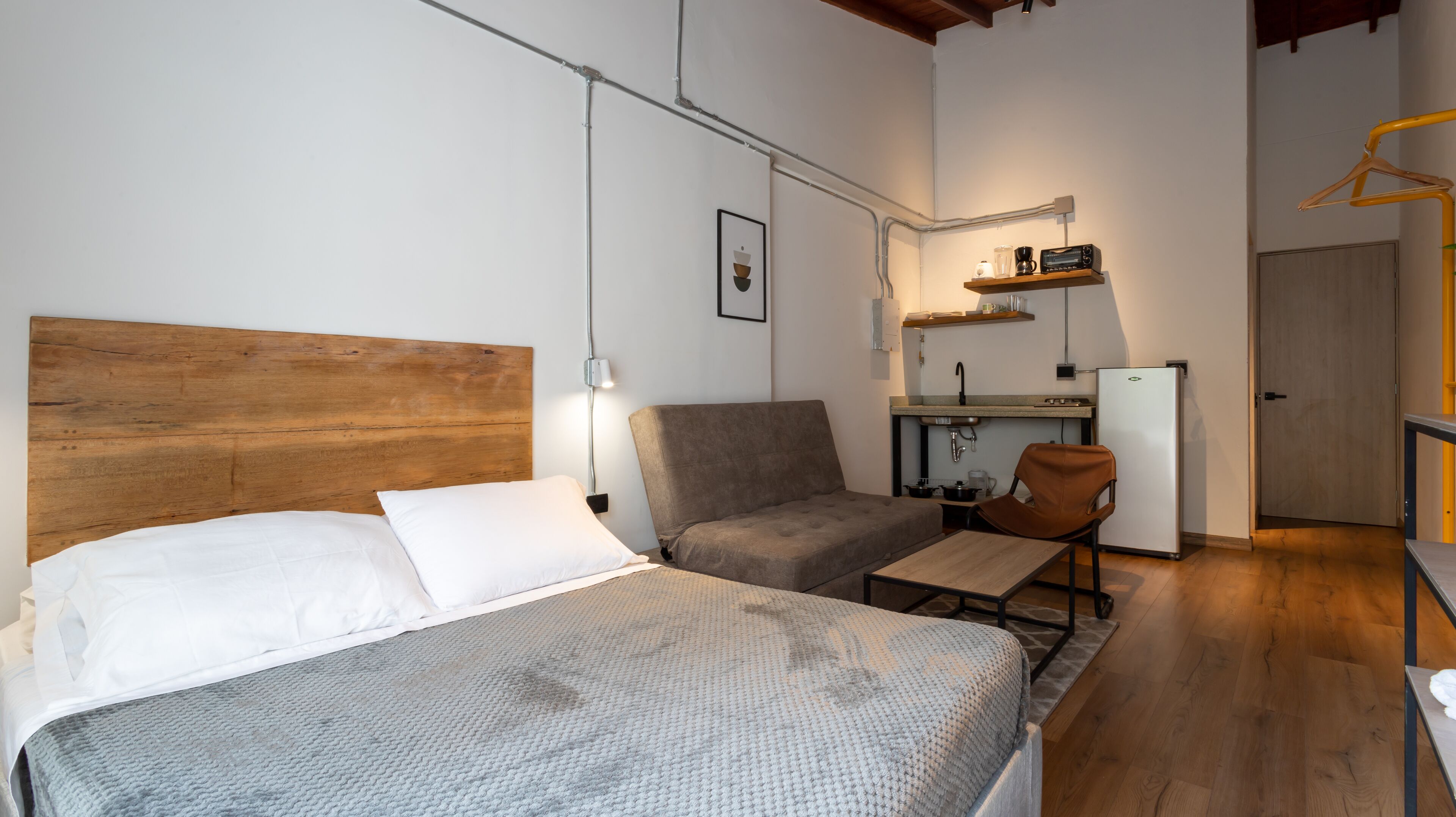 Una camera da letto compatta con una testiera in legno, un letto con lenzuola bianche, un divano grigio, un piccolo tavolo e una sedia. Si intravede sullo sfondo un angolo cottura con un lavello e un frigorifero.
