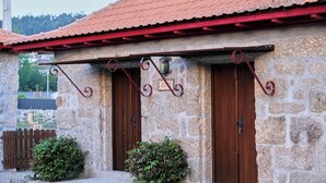 A stone building with a red-tiled roof, two wooden doors, and a small garden area with bushes.