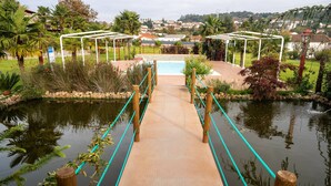 A wooden bridge over a pond with a pool in the background, surrounded by greenery and a view of houses.