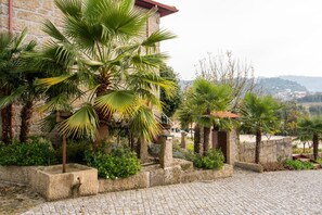 Une cour pavée de pierres, agrémentée de palmiers, bordée de murs en pierre et offrant une vue sur une ville lointaine.