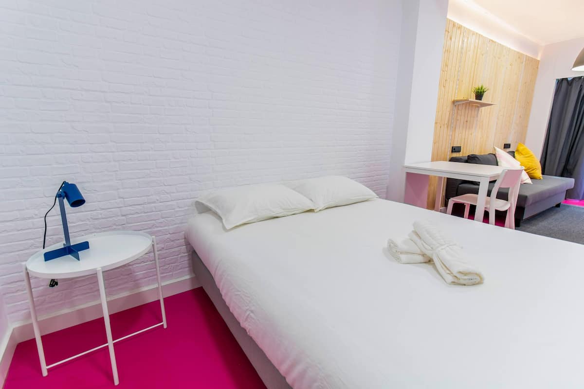 Une chambre d’hôtel avec un mur en briques blanches, un lit bien fait avec une literie blanche, une petite table ronde avec une lampe, et un coin salon avec un canapé et un fauteuil.
