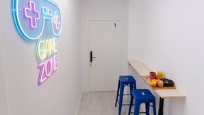 Um quarto com um letreiro de neon que diz "GAME ZONE", uma porta com a etiqueta "1", uma mesa de madeira com uma bandeja de comida e bebidas, e dois bancos azuis.