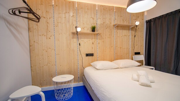 Quarto de hotel moderno com painel de parede em madeira, cama com roupa de cama branca, mesa redonda pequena e luminária pendente.