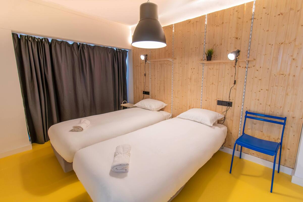 Une chambre avec deux lits, une chaise bleue, une lampe suspendue et une étagère avec une plante.