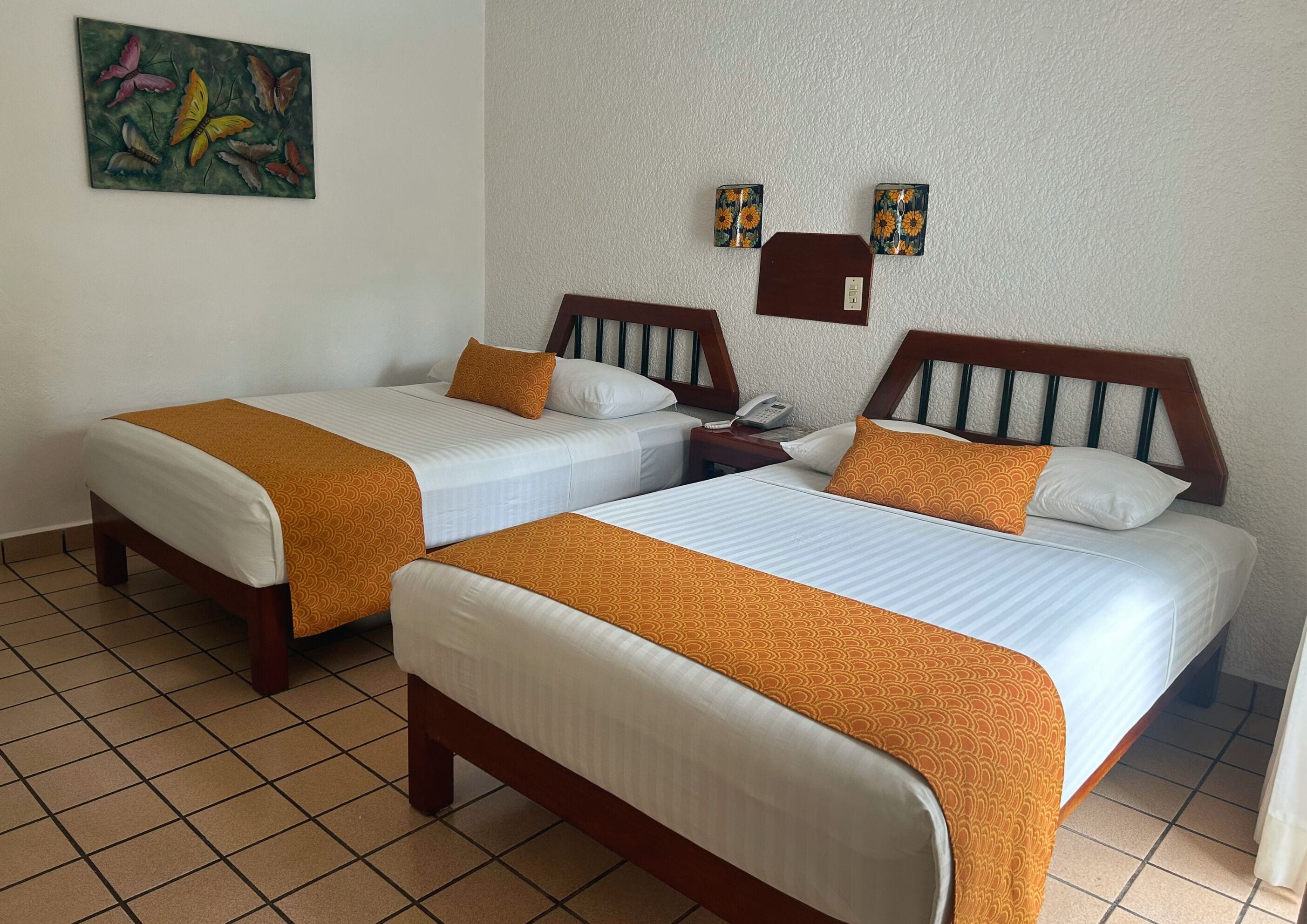 Habitación de hotel con dos camas, cada una con cabecero de madera, ropa de cama blanca y almohadas decorativas naranjas. Hay una mesita de noche con un teléfono y una pequeña mesa con una lámpara. Un cuadro cuelga en la pared sobre las camas.