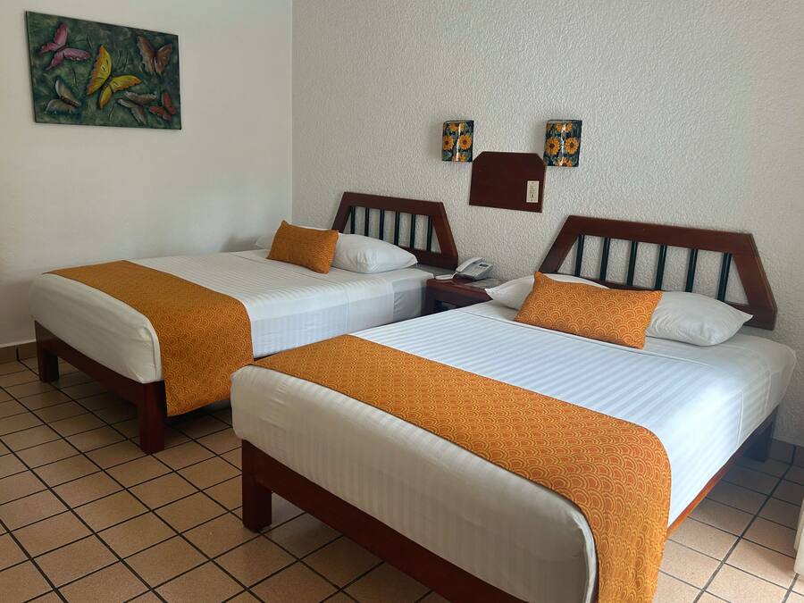 Habitación de hotel con dos camas, cada una con cabecero de madera, ropa de cama blanca y almohadas decorativas naranjas. Hay una mesita de noche con un teléfono y una pequeña mesa con una lámpara. Un cuadro cuelga en la pared sobre las camas.