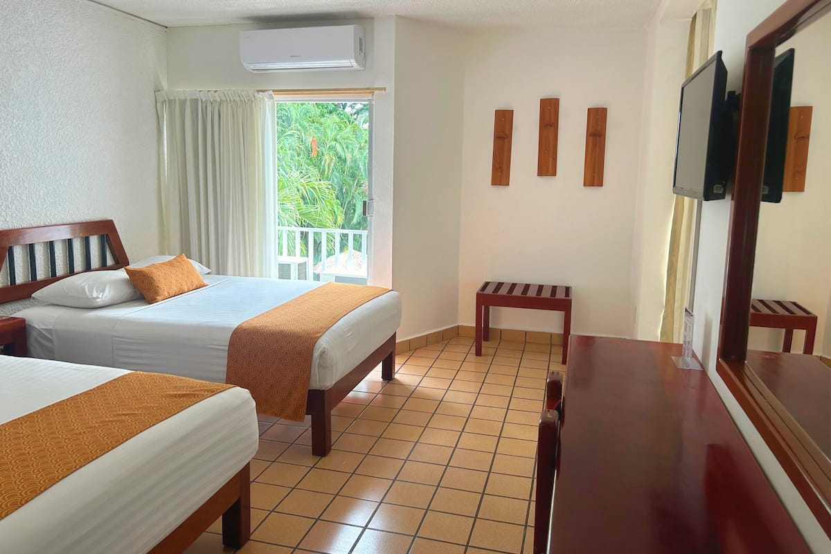 Habitación de hotel con dos camas, cabecero de madera, una mesita, televisor y balcón con cortinas.