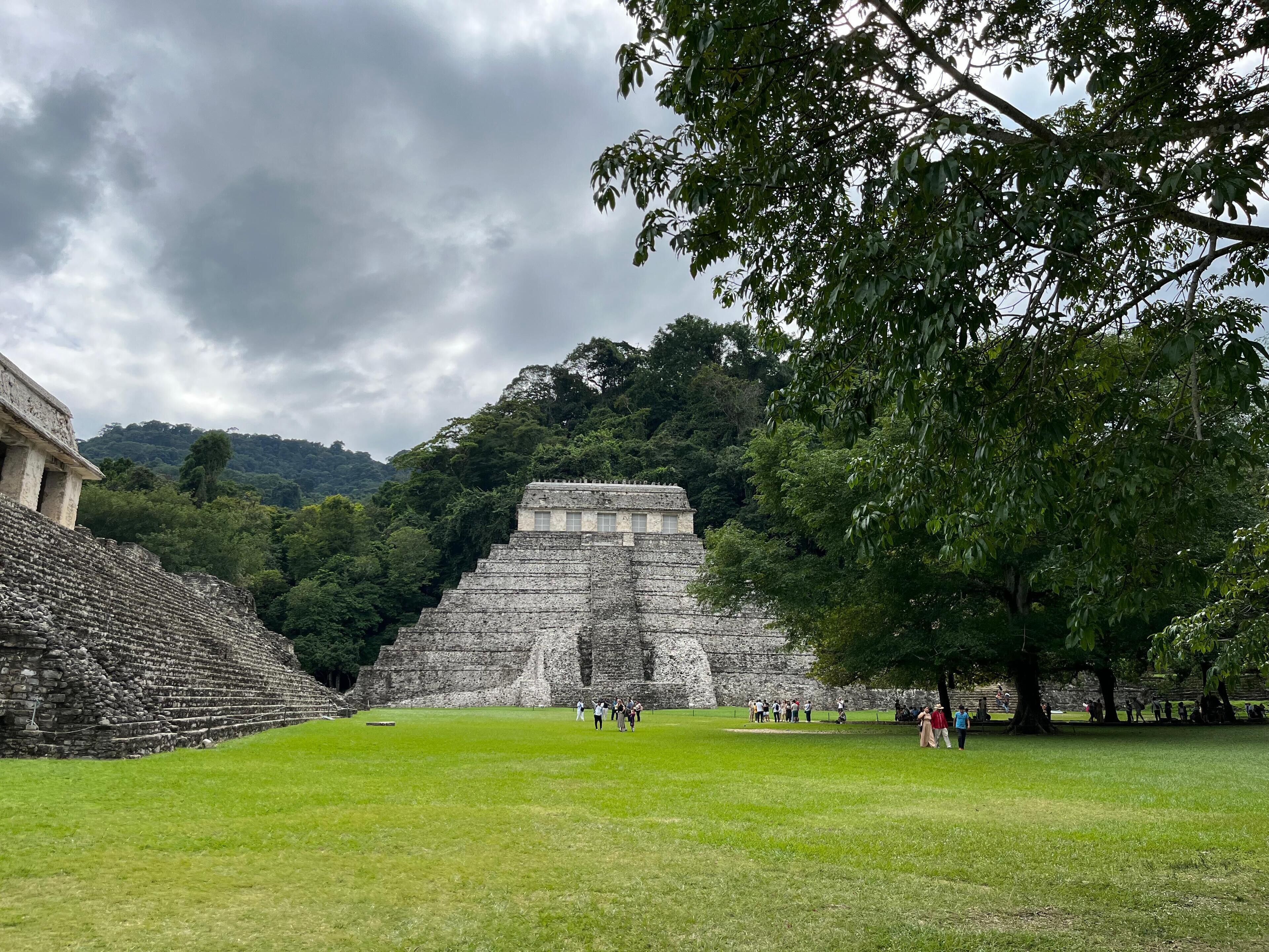 A large ancient pyramid structure surrounded by lush greenery and a cloudy sky.