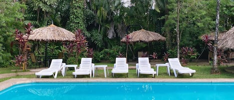 A poolside area with white lounge chairs and thatched umbrellas, surrounded by lush greenery.