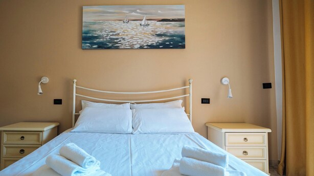 A neatly made bed with white linens and towels, two bedside tables with lamps, a painting of sailboats on the wall, and a beige wall color. - Marina Bike Family Home (Andora)