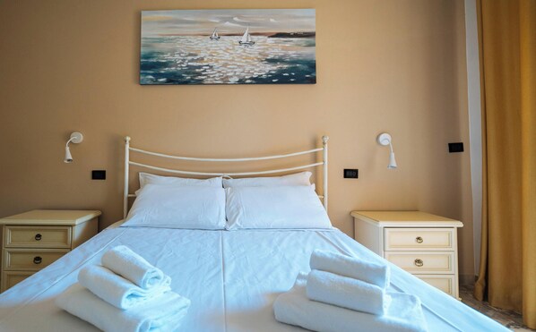 A neatly made bed with white linens and towels, two bedside tables with lamps, a painting of sailboats on the wall, and a beige wall color. - Marina Bike Family Home (Andora)