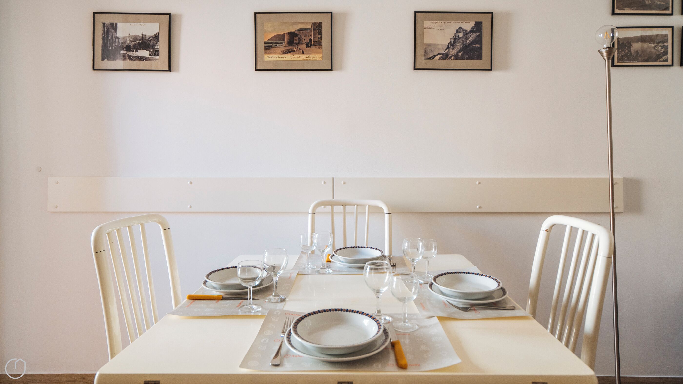 Mesa de jantar posta com pratos, copos e talheres, acompanhada por cadeiras brancas, com quadros na parede acima.
