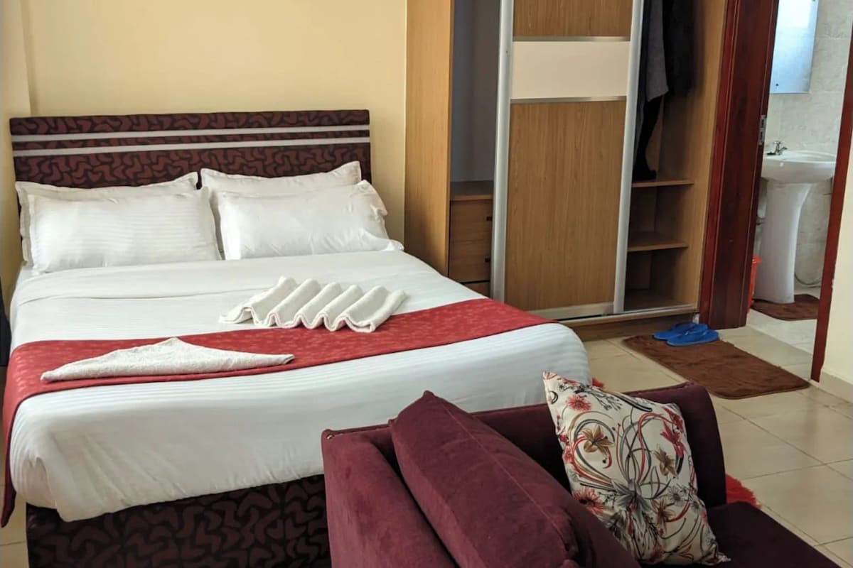 Quarto de hotel com cama, guarda-roupa, sofá e banheiro visível através de uma porta aberta.