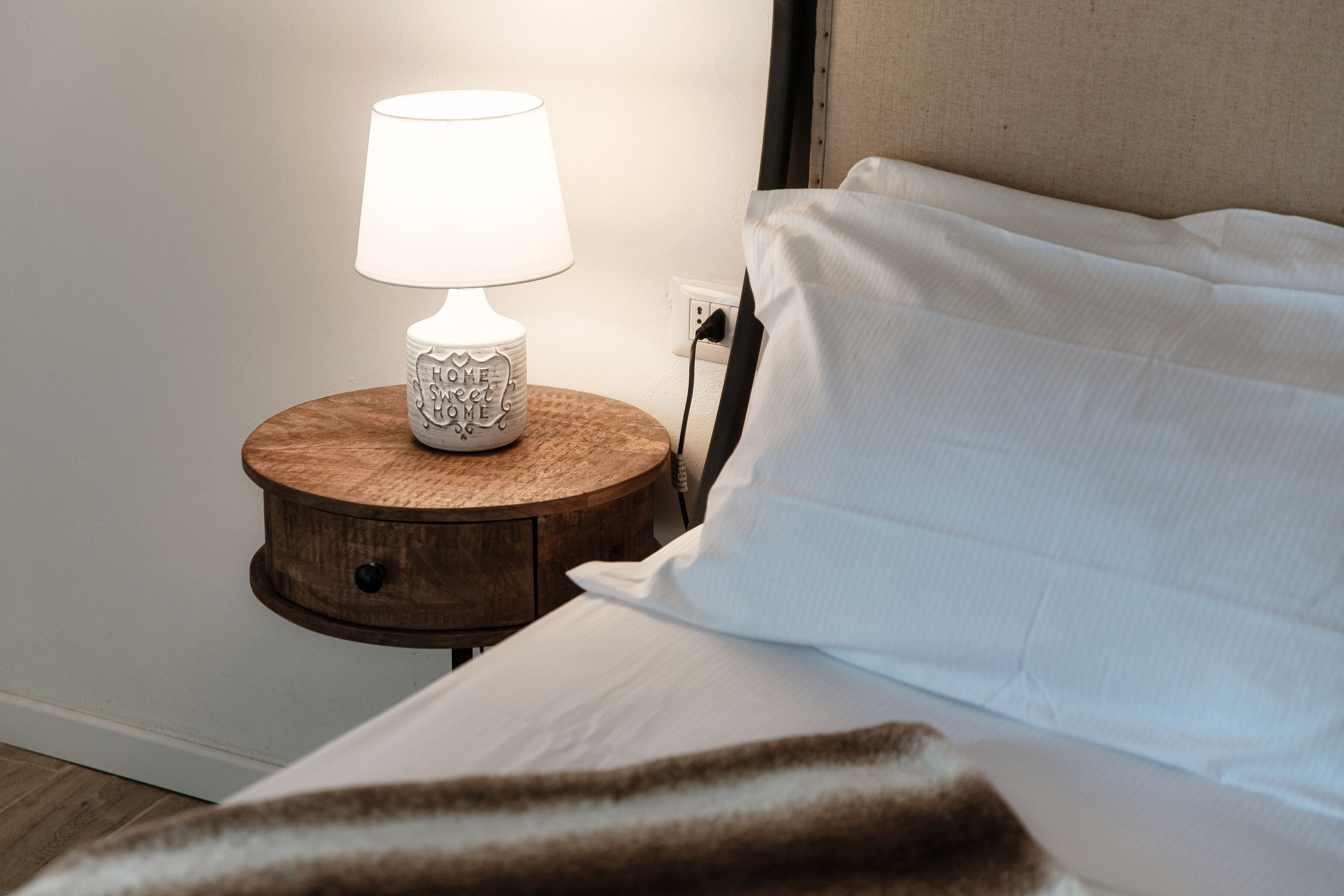 Ein ordentlich bezogenes Bett mit weißen Laken und einer braunen Decke, ein runder Nachttisch mit einer weißen Lampe sowie ein beige farbener Kopfteil.