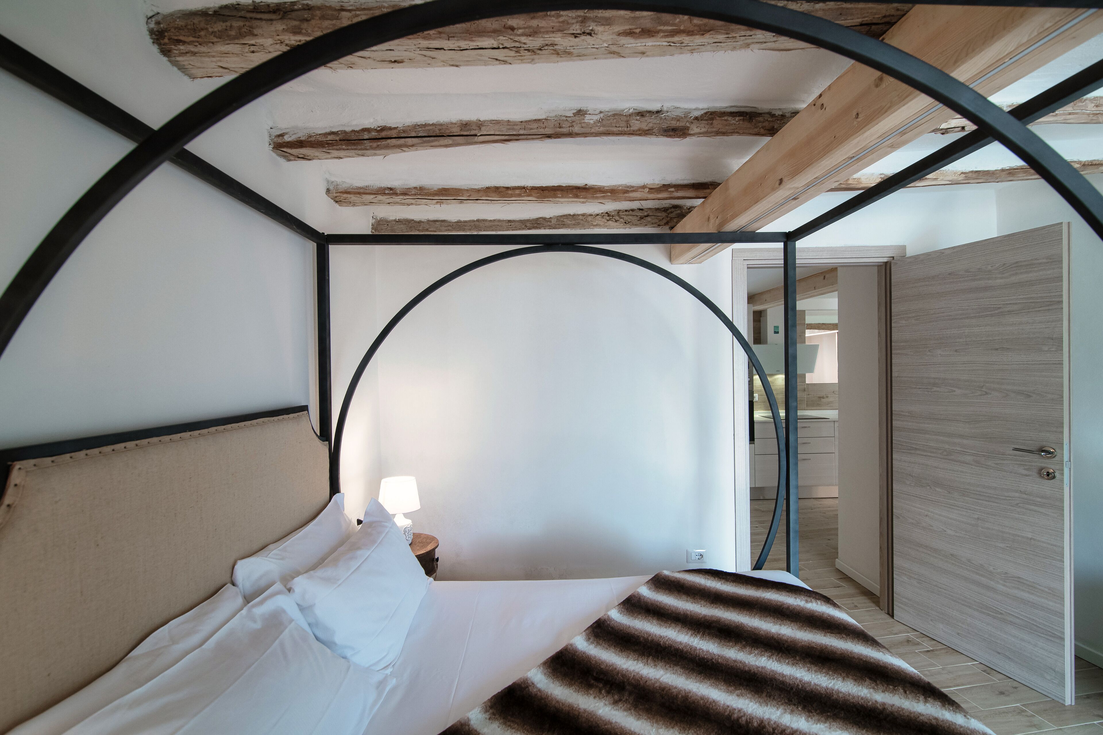 A bed with white linens and a striped blanket, a nightstand with a lamp, and a wooden ceiling with exposed beams.