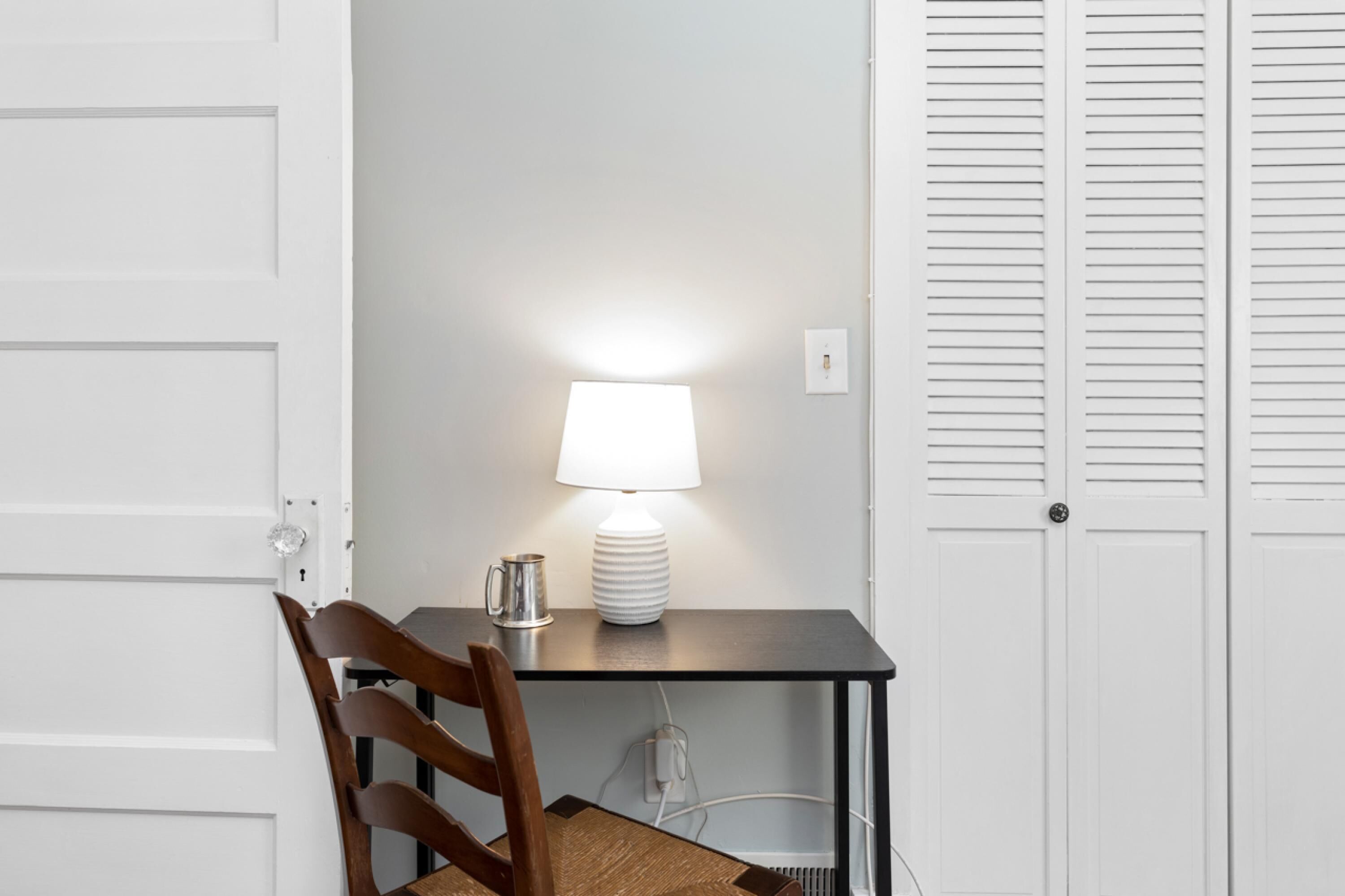 A small, dark-colored desk with a white lamp, a wooden chair, and a white door with louvered panels.