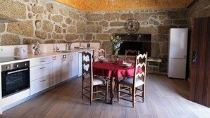 A kitchen with stone walls, wooden ceiling beams, a chandelier, a dining table with chairs, and a refrigerator. - Aldeia da Quinta do Paço- Casa da Criada (São Salvador do Campo)