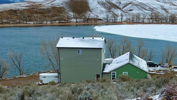 Une maison verte, dont le toit est recouvert de neige, située près d’un lac, avec des montagnes en arrière-plan.