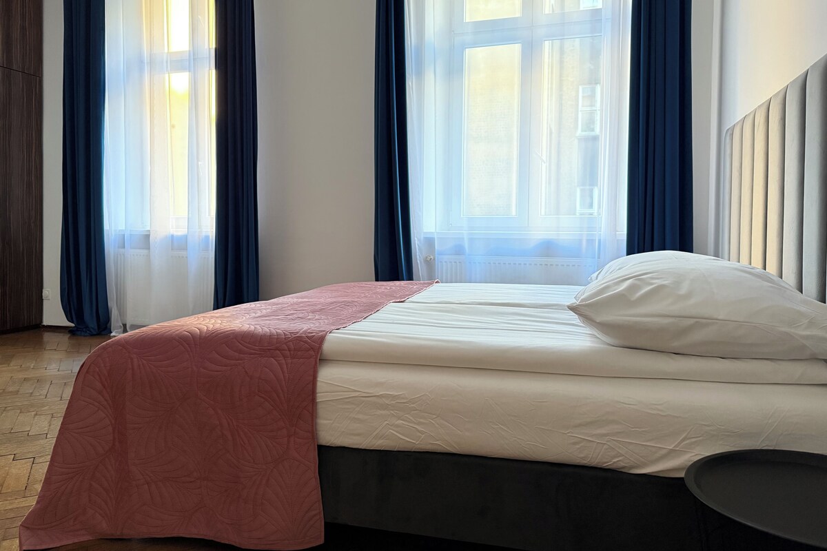 A neatly made bed with a pink blanket and white pillows, a dark headboard, and a small black side table.