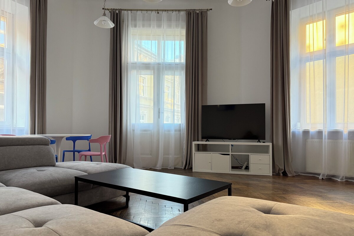 Un salon moderne avec un canapé, une table basse, un téléviseur posé sur un meuble blanc et une vue sur les immeubles à travers de grandes fenêtres.