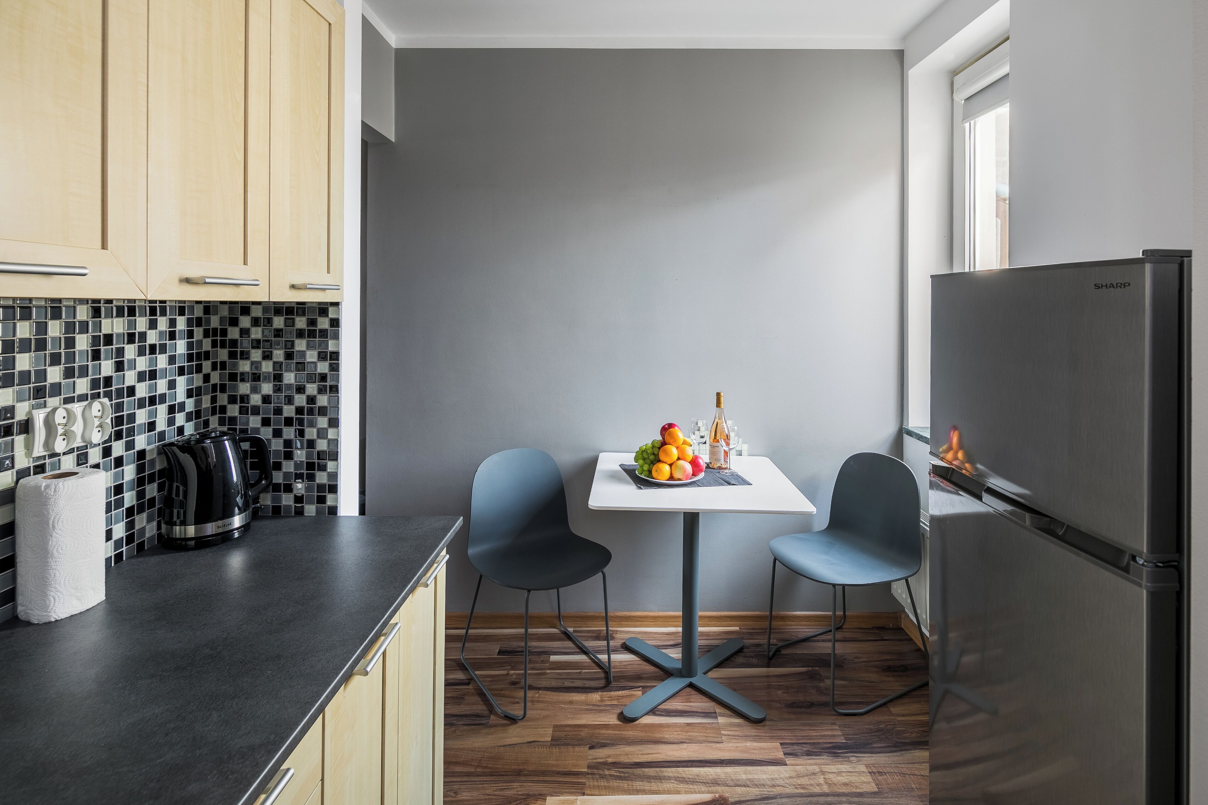 Une cuisine compacte avec un plan de travail noir, deux chaises, une petite table avec des fruits et une bouteille, et un réfrigérateur.
