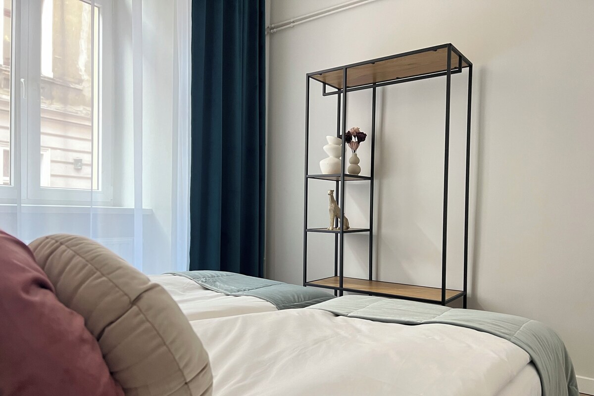 Un dormitorio con una cama, una ventana con cortinas y una estantería metálica con objetos decorativos.