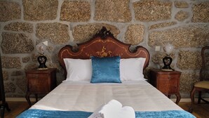 Een net opgemaakt bed met een blauwe deken en een opgerolde handdoek, een houten hoofdbord met gouden details, en muren met een stenen patroon.