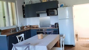 A modern kitchen with dark cabinets, a microwave, a stove, and a refrigerator. There is a dining table with chairs and a hanging light fixture above it. - Jolie Maison Entre Mer et Montagne (Brando)