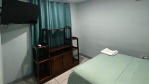 Una habitación con cama, una silla de madera, un televisor montado en la pared y un equipo de aire acondicionado.