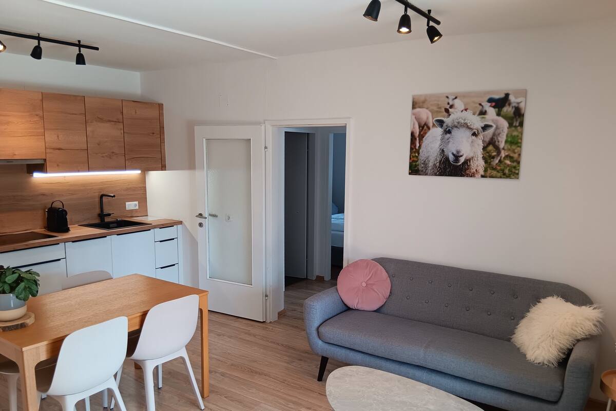 A modern kitchen with wooden cabinets, a dining table with chairs, a grey sofa with a pink pillow, and a small round table.