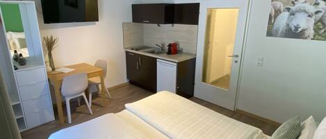 A hotel room with a bed, a desk, a kitchenette, and a door with a mirror.