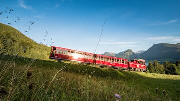 Un tren rojo que avanza por un paisaje montañoso con abundante vegetación y cielos despejados.