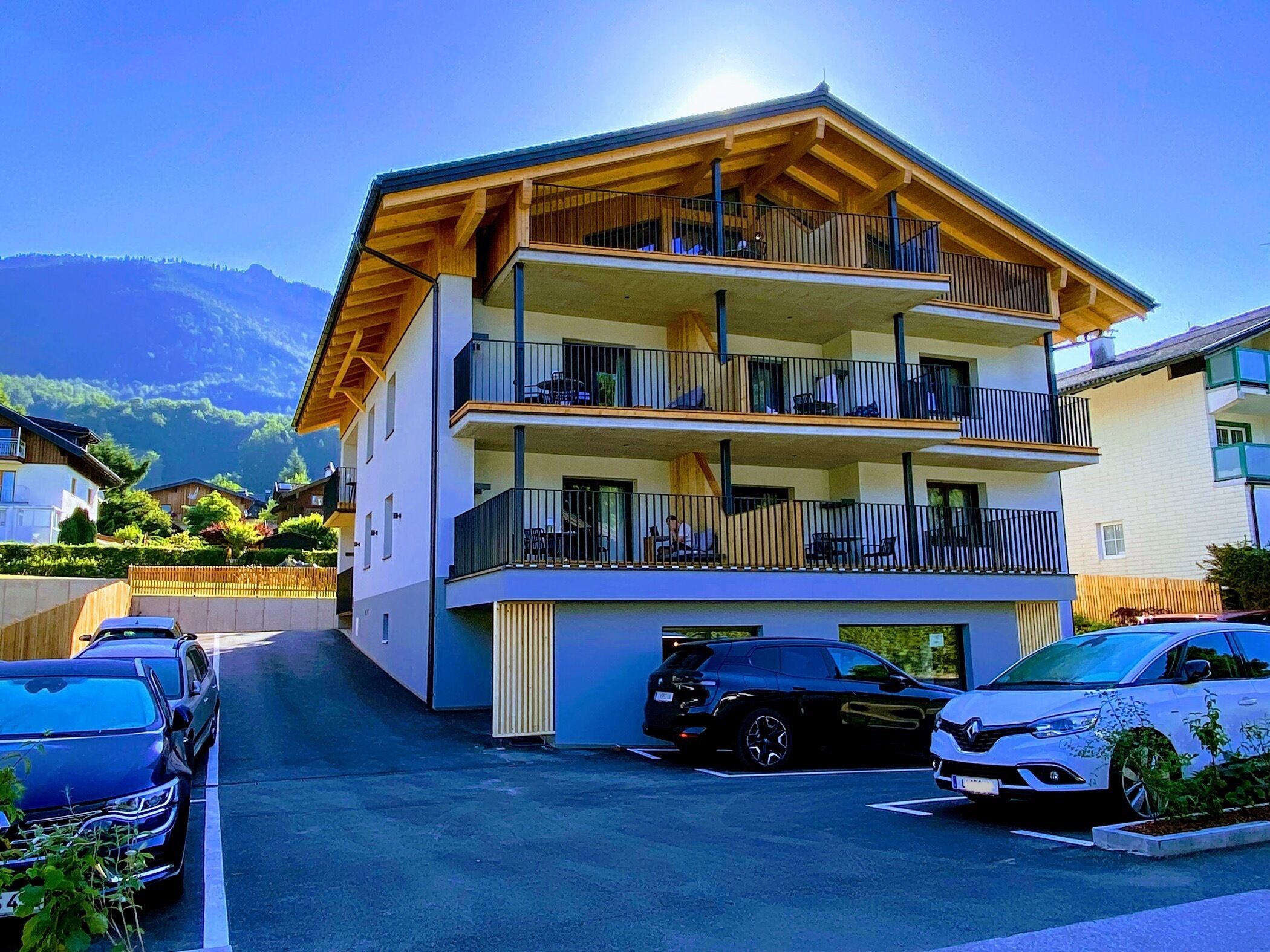 A multi-story building with balconies, a parking area with cars, and mountains in the background.
