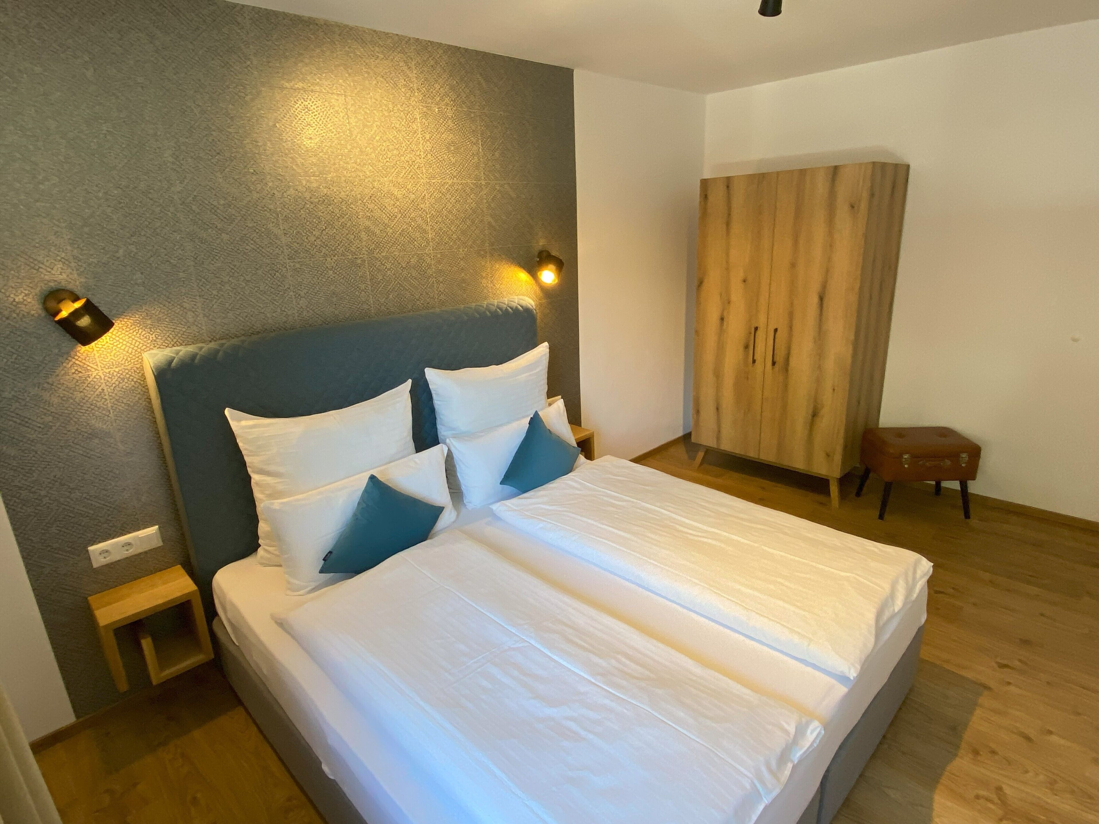 A neatly made bed with white and blue pillows, a wooden wardrobe, and a small wooden stool in a modern room with a textured wall and a wall-mounted light fixture.