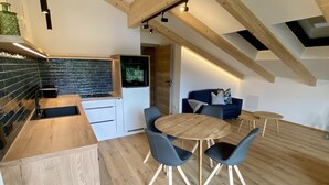 A modern kitchen with a built-in oven, a dining area with a round wooden table and chairs, and a living space with a sofa and a TV. - Wolf & Schaf Apartments (St. Gilgen)