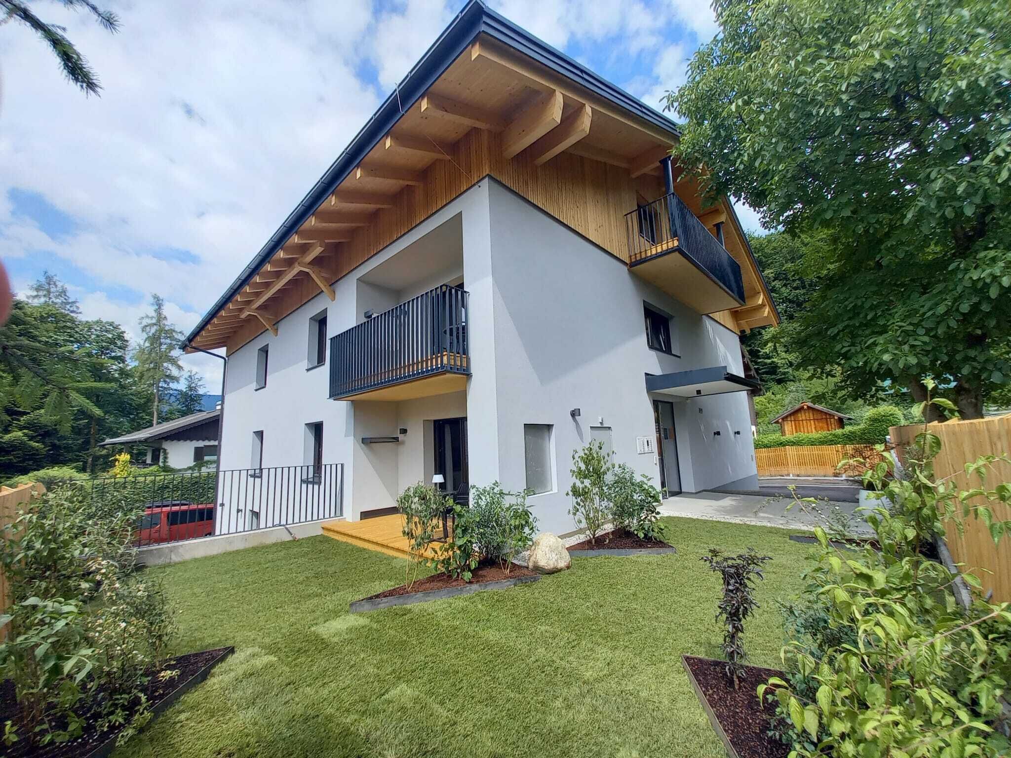 Une maison moderne à deux étages, dotée d’un balcon, entourée d’une pelouse bien entretenue et de diverses plantes.