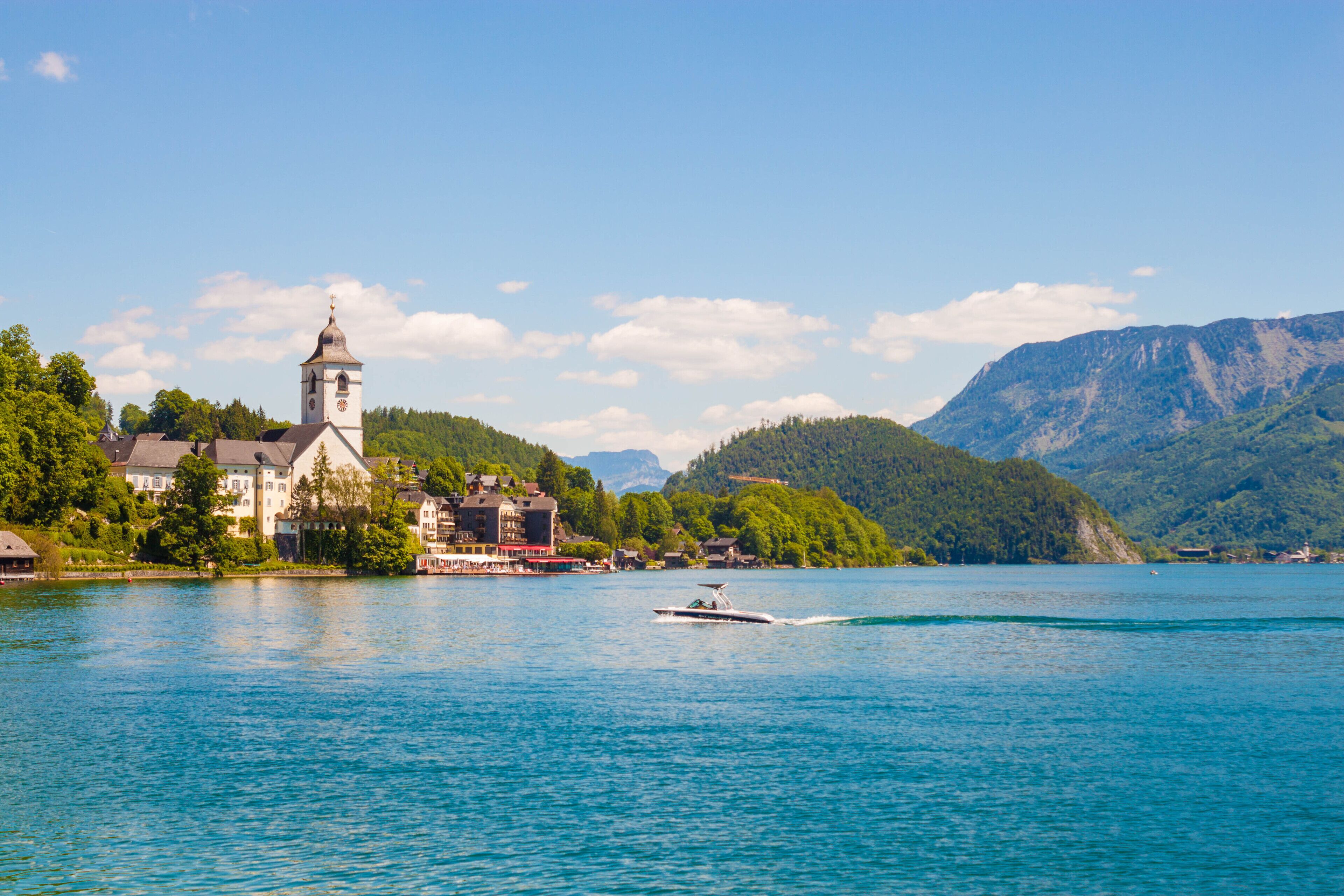 Eine Stadt am Seeufer mit einem markanten Kirchengebäude, einem Boot auf dem Wasser und Bergen im Hintergrund.