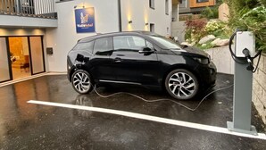 A black car parked in a covered parking area with a charging station, a building with a sign for 'WolfSchaft Apartments', and a balcony with a railing. - Wolf & Schaf Apartments (St. Gilgen)