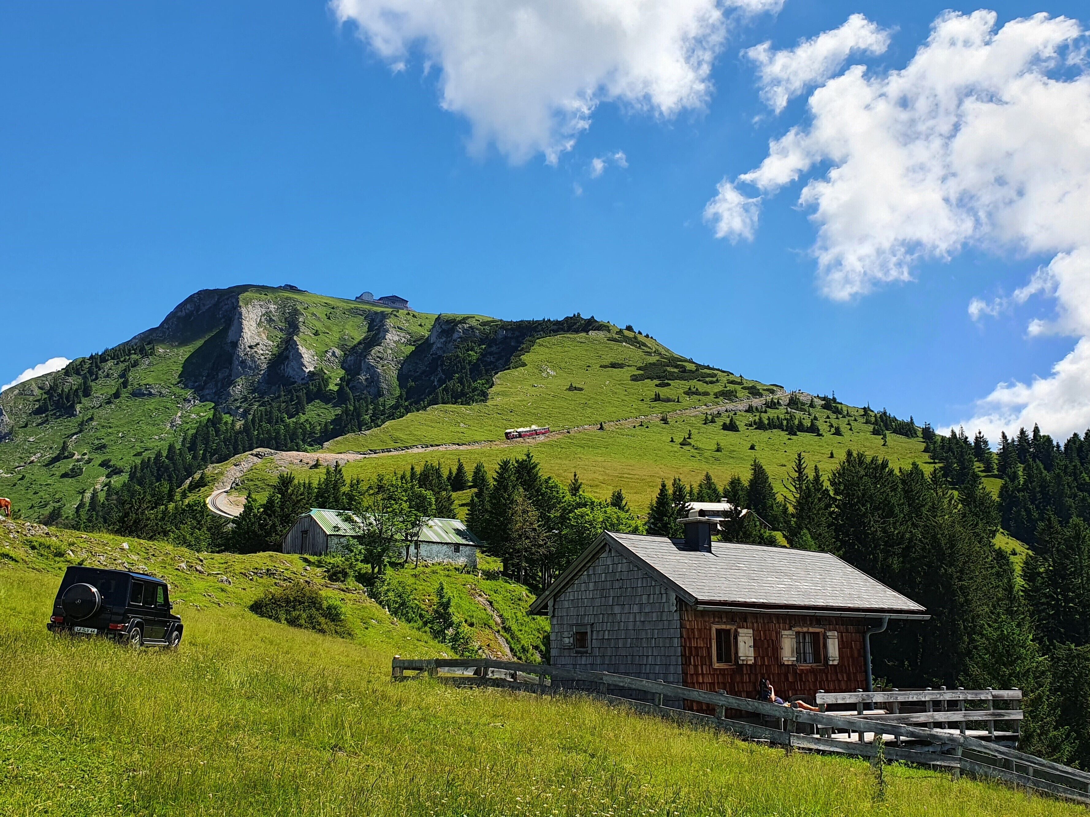 A rural landscape with a mountain, a small wooden cabin, and a parked car.