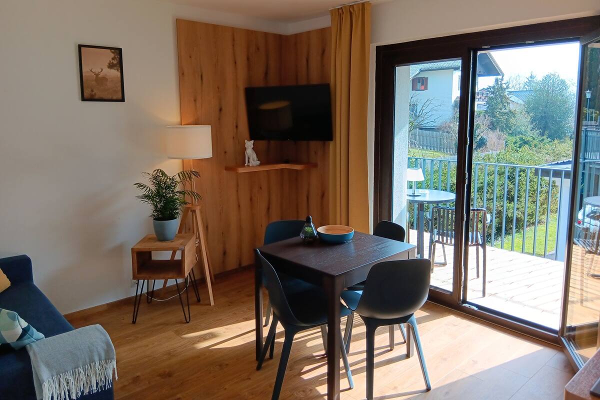 A modern living room with a wooden accent wall, a flat-screen TV, a dining table with chairs, a sofa, and a large sliding glass door leading to a balcony with outdoor furniture.
