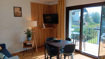 Una sala de estar moderna con una pared de acento de madera, un televisor de pantalla plana, una mesa de comedor con sillas, un sofá y un amplio ventanal corredera que da a un balcón con mobiliario exterior.