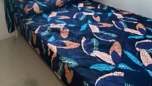 Een eenpersoonsbed met een sprei met bladmotief in een kamer met een raam, gordijnen en een aan de muur hangend kunstwerk.
