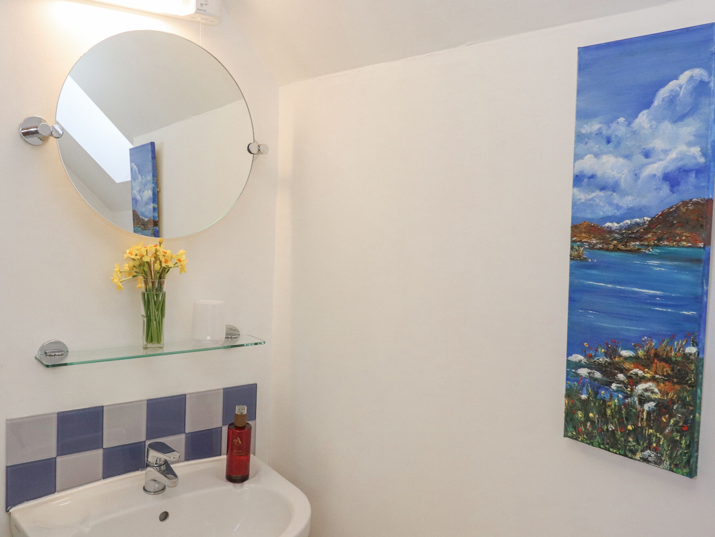 A bathroom with a round mirror, a glass shelf with flowers, a sink, and a painting of a landscape.