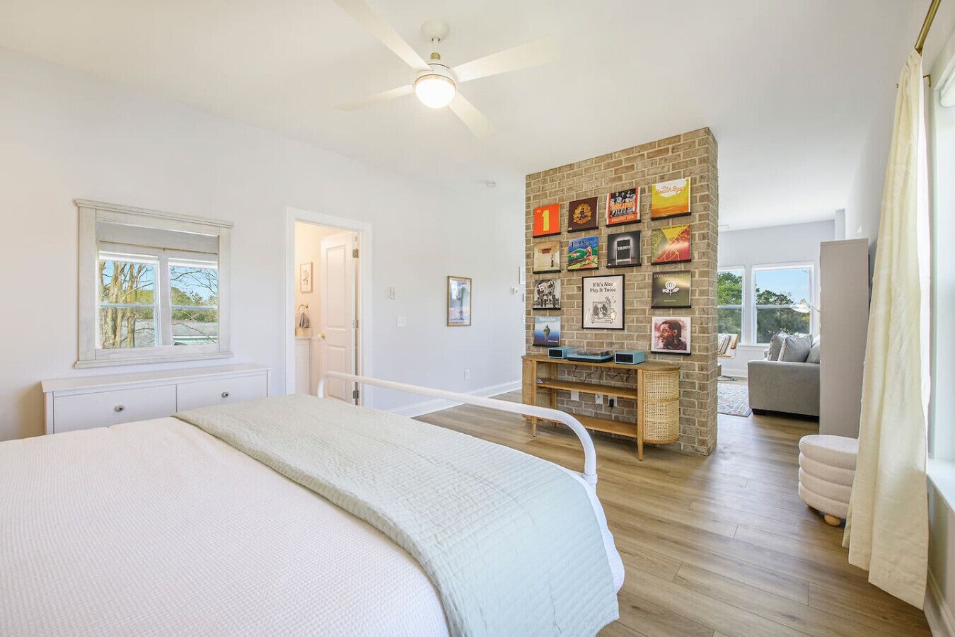 A bedroom with a bed, a ceiling fan, a brick accent wall with framed pictures, a window, and a door.