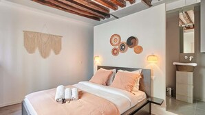 A modern bedroom with a wooden ceiling, a bed with pink bedding, a wall-mounted macrame decoration, and a bathroom visible through an open door.
