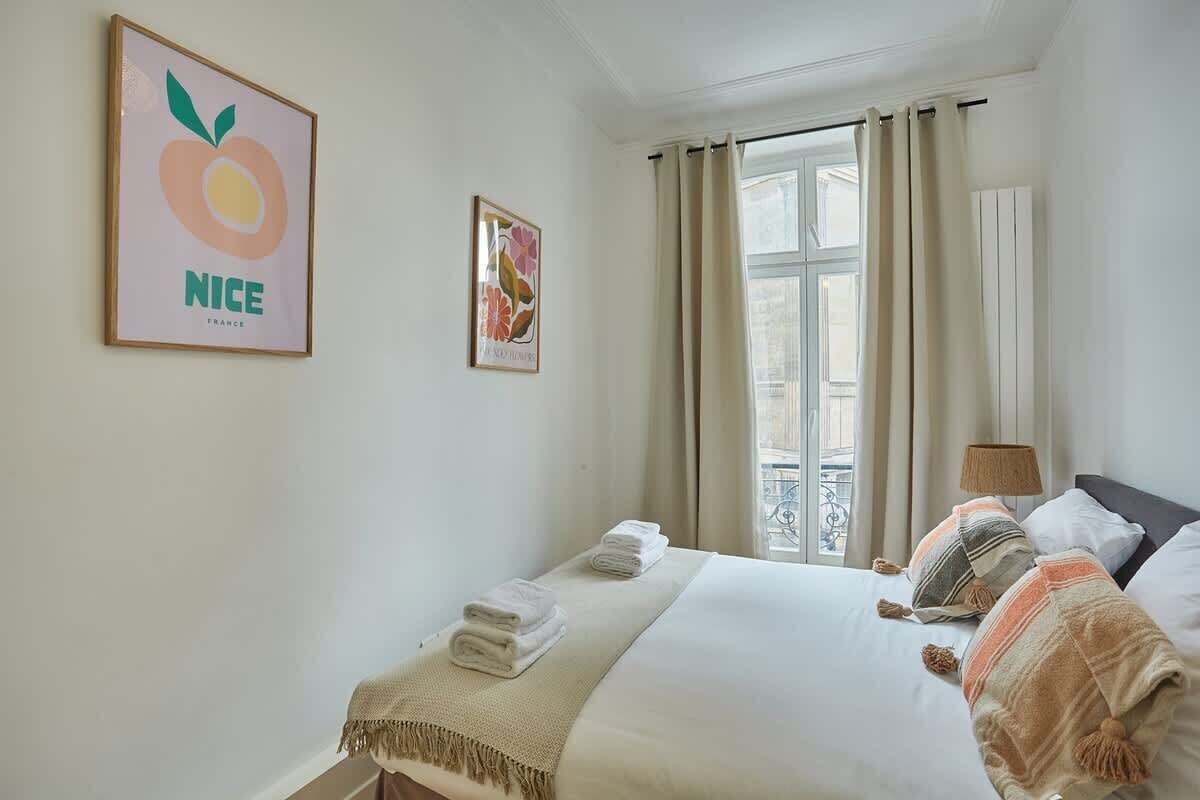 Une chambre d’hôtel comprenant un lit, deux oreillers, une lampe de chevet, une fenêtre avec des rideaux et deux œuvres d’art murales.