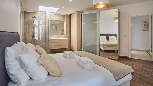 A modern bedroom with a large bed, a walk-in closet, a glass shower enclosure, and a view of a cityscape.