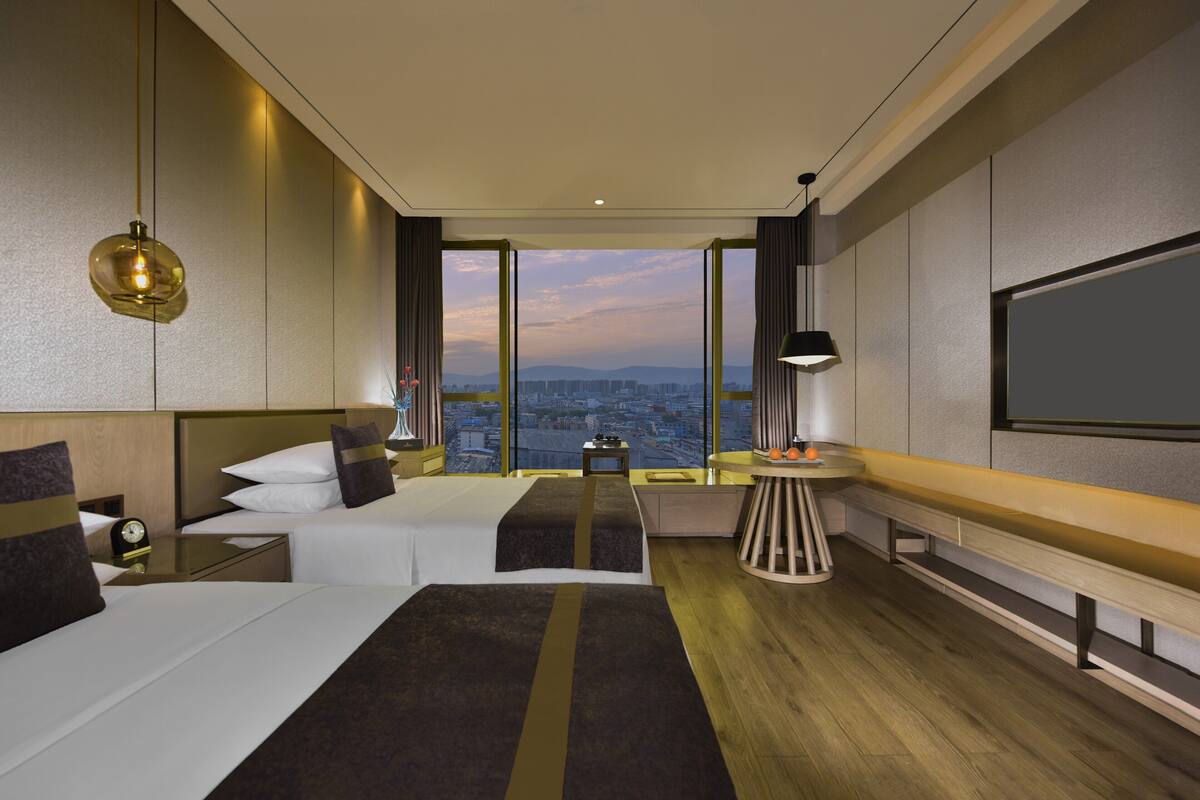 A modern hotel room with a large bed, a desk, a small table, and a large window offering a city view.