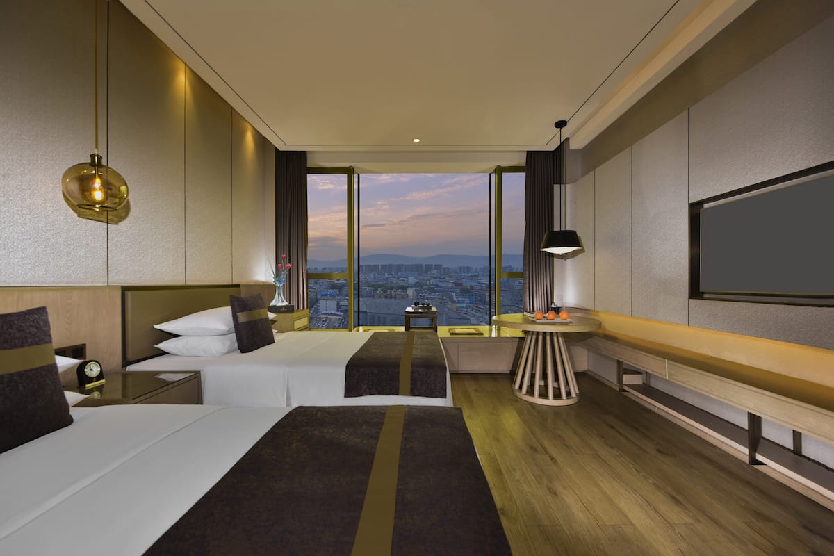 A modern hotel room with a large bed, a desk, a small table, and a large window offering a city view.